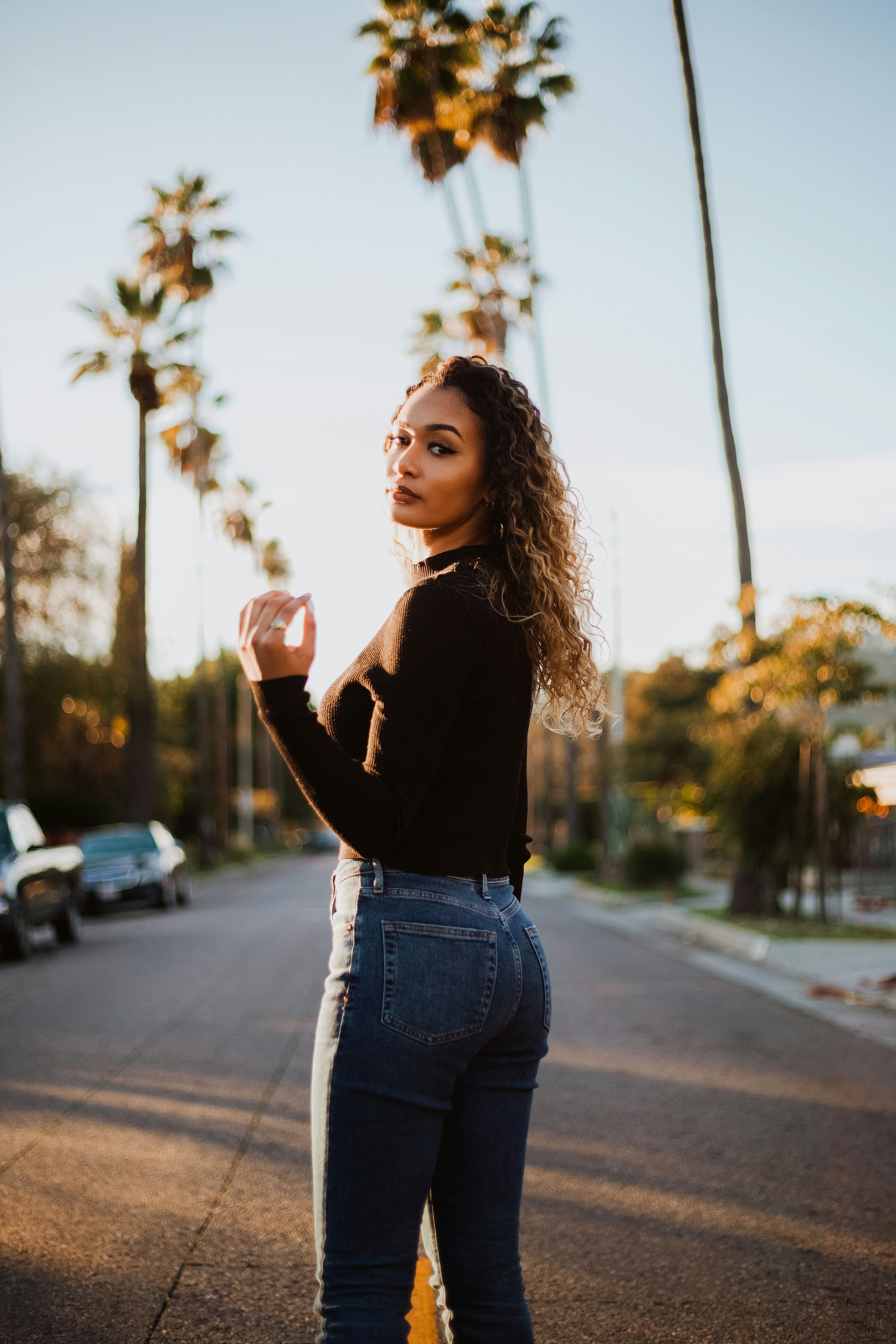 woman standing in the middle of the road while looking over her left shoulder near parked cars and trees during day